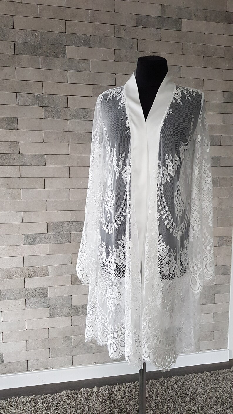 May include: White lace bridal robe with a satin lining. The robe has long sleeves and a relaxed fit.  Delicate floral lace patterns adorn the sheer fabric. The robe is displayed on a mannequin against a textured white brick wall. This elegant, lightweight garment is perfect for bridal preparations or special occasions.
