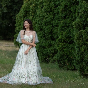 May include: A woman in a white dress with floral embroidery stands in a grassy field. The dress has a sheer overlay and a cape-like top. She is smiling and looking to the side.