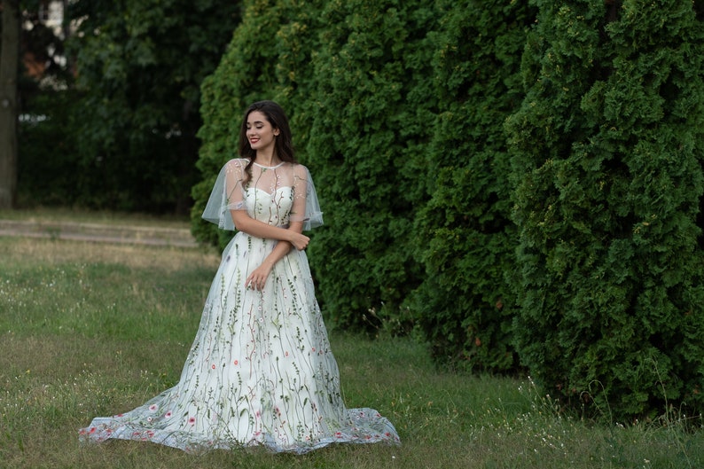 May include: A woman in a white dress with floral embroidery stands in a grassy field. The dress has a sheer overlay and a cape-like top. She is smiling and looking to the side.