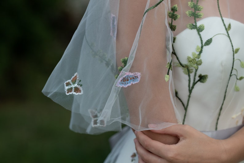 May include: A white dress with a sheer overlay featuring embroidered butterflies and floral details.