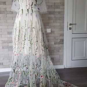 May include: A white tulle gown with floral embroidery. The gown has a fitted bodice and a flowing skirt. The embroidery features delicate flowers and leaves in shades of pink, green, and red.
