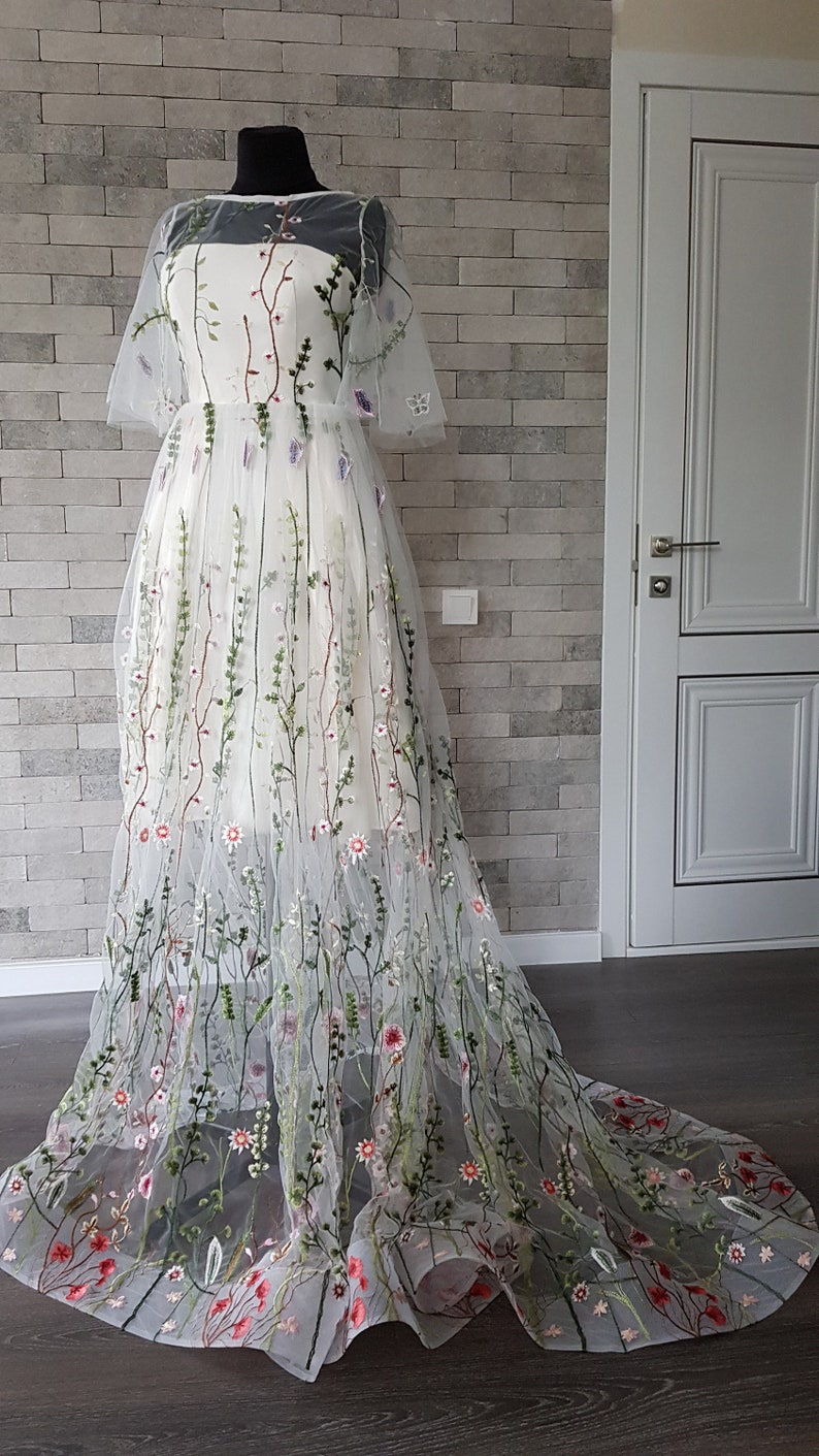 May include: A white tulle gown with floral embroidery. The gown has a fitted bodice and a flowing skirt. The embroidery features delicate flowers and leaves in shades of pink, green, and red.