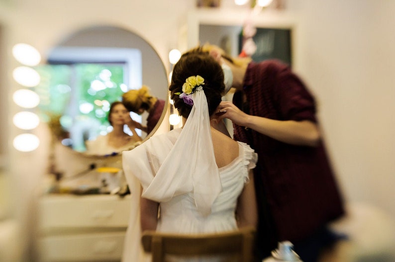 Op de afbeelding: Een bruid met een witte sluier en bloemen in haar haar wordt gecoiffeerd door een styliste. De bruid zit op een stoel en de styliste staat achter haar.