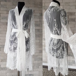 May include: White lace bridal robe with long sleeves and a satin sash. The robe is shown in two views: front and back.  Delicate floral lace patterns adorn the sheer fabric. The robe features a classic kimono style with a self-tie belt.  It's a luxurious and elegant garment perfect for bridal preparations or special occasions.