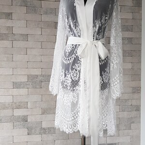 May include: A white lace bridal robe with a satin sash. The robe has long sleeves and a knee-length hem. The lace is delicate and features floral patterns. The satin sash is tied at the waist. The robe is displayed on a mannequin against a brick wall background.