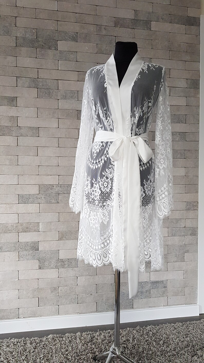May include: A white lace bridal robe with a satin sash. The robe has long sleeves and a knee-length hem. The lace is delicate and features floral patterns. The satin sash is tied at the waist. The robe is displayed on a mannequin against a brick wall background.