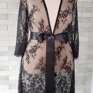May include: Black lace robe with a satin sash. The robe is sheer with a floral pattern and has three-quarter length sleeves.  The robe is knee-length and has a relaxed fit.  It is displayed on a mannequin against a textured gray background.