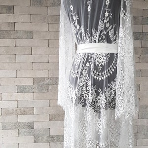 May include: White lace bridal robe with long sleeves and a satin sash. The robe has a scalloped hemline and delicate floral embroidery.  The sheer lace fabric creates an elegant and romantic look, perfect for bridal preparations or a luxurious boudoir experience.  This romantic robe is ideal for getting ready for a wedding or special occasion.