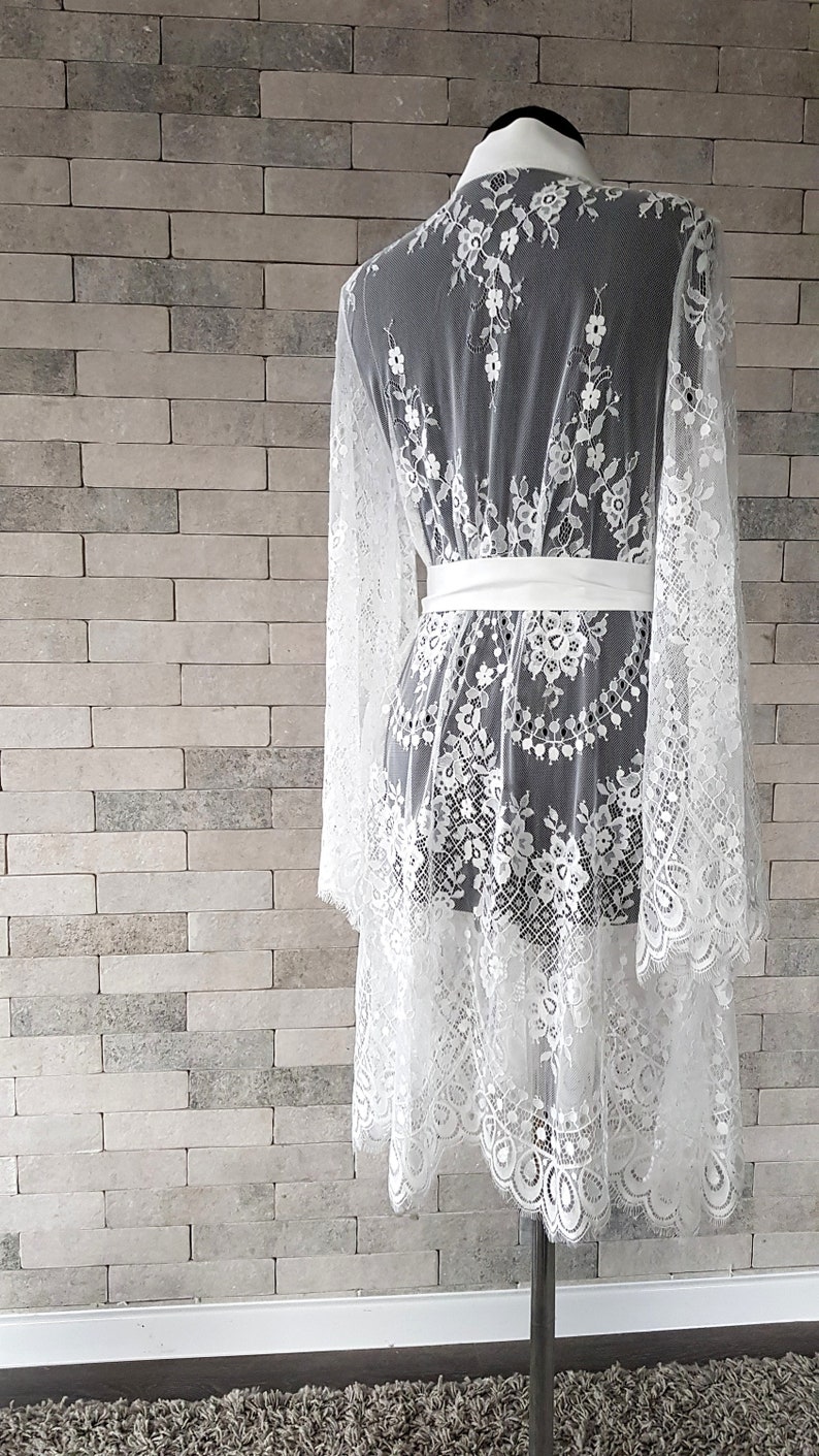 May include: White lace bridal robe with long sleeves and a satin sash. The robe has a scalloped hemline and delicate floral embroidery.  The sheer lace fabric creates an elegant and romantic look, perfect for bridal preparations or a luxurious boudoir experience.  This romantic robe is ideal for getting ready for a wedding or special occasion.