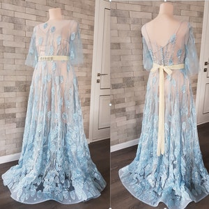 May include: A light blue, floor-length gown with embroidered floral details. The dress features a sheer overlay, creating a delicate and romantic look.  It has a fitted bodice and a flowing skirt.  A pale yellow sash is tied around the waist. The dress is shown from the front and back on dress forms.