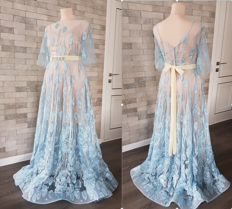 May include: A light blue, floor-length gown with embroidered floral details. The dress features a sheer overlay, creating a delicate and romantic look.  It has a fitted bodice and a flowing skirt.  A pale yellow sash is tied around the waist. The dress is shown from the front and back on dress forms.