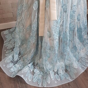May include: A light blue maxi dress with embroidered floral details. The dress features a sheer, flowing fabric with delicate, light blue floral embroidery. A satin ribbon is tied around the waist. The hemline is scalloped and features a subtle train. The dress is displayed on a wooden floor.