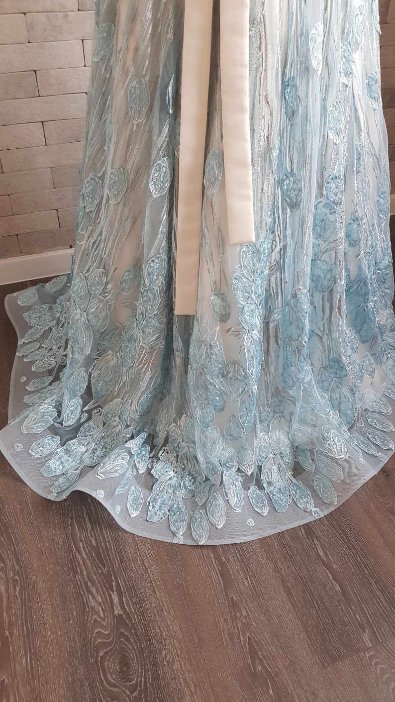 May include: A light blue maxi dress with embroidered floral details. The dress features a sheer, flowing fabric with delicate, light blue floral embroidery. A satin ribbon is tied around the waist. The hemline is scalloped and features a subtle train. The dress is displayed on a wooden floor.
