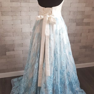 May include: A light blue and white a-line gown with embroidered floral details. The dress features a sweetheart neckline, a fitted bodice, and a flowing skirt.  A white satin sash is tied at the waist. The back of the dress is open. This elegant formal gown is perfect for a special occasion.