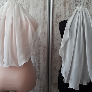 Op de afbeelding: Twee witte bruids sluiers tentoongesteld op mannequins. De sluiers zijn gemaakt van zachte, doorschijnende stof en hebben een delicaat, vloeiende ontwerp.