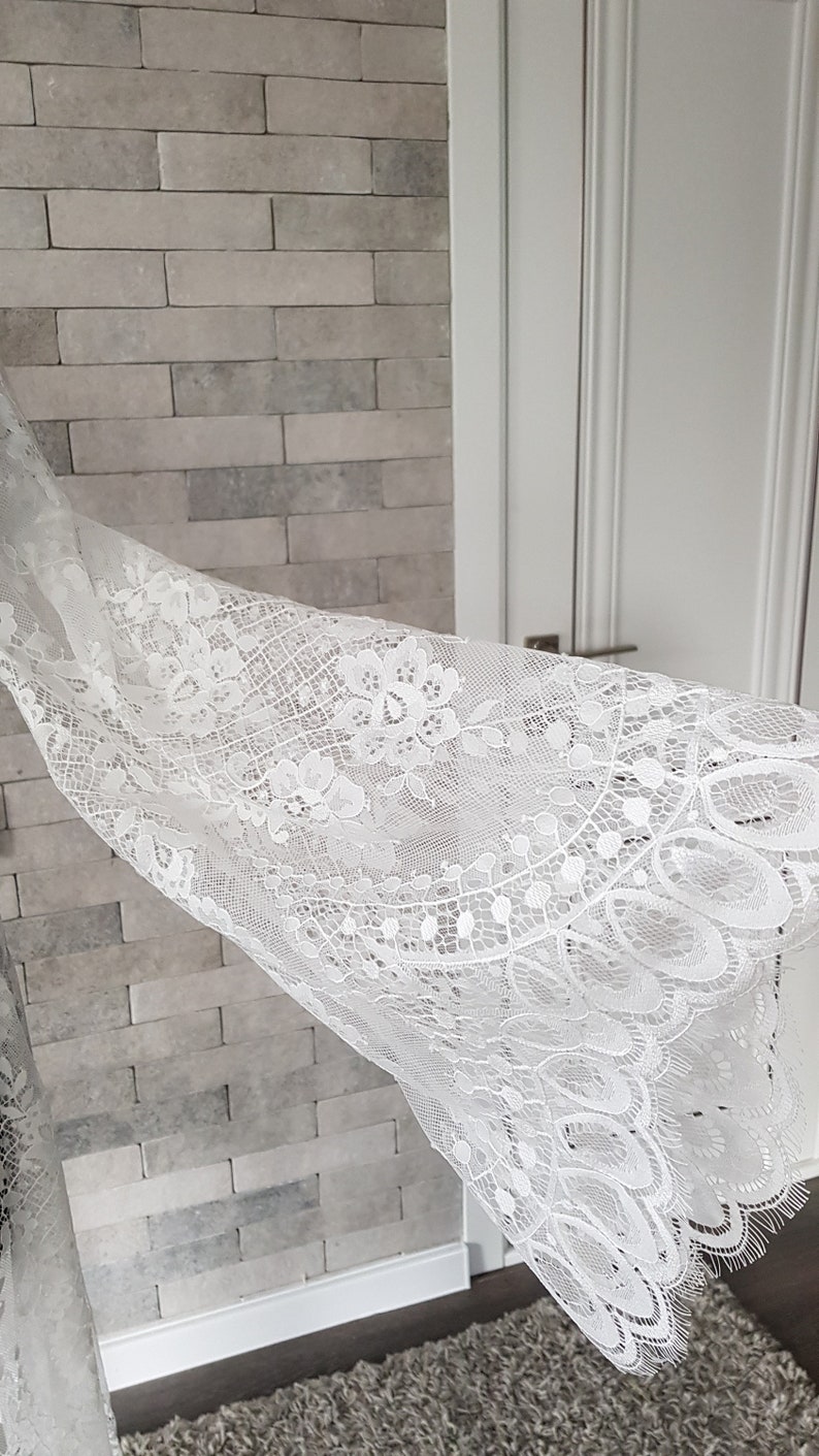 May include: Close-up of white lace fabric with a floral pattern. The lace has scalloped edges and delicate detailing. The fabric is shown draped over a textured gray brick wall.  The lace is sheer and lightweight, with intricate floral designs throughout.  It appears to be a bridal or special occasion fabric.