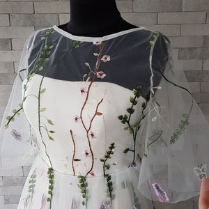 May include: A white tulle dress with floral embroidery. The dress has a sheer overlay with a round neckline and short sleeves. The embroidery features green leaves, pink flowers, and brown stems.