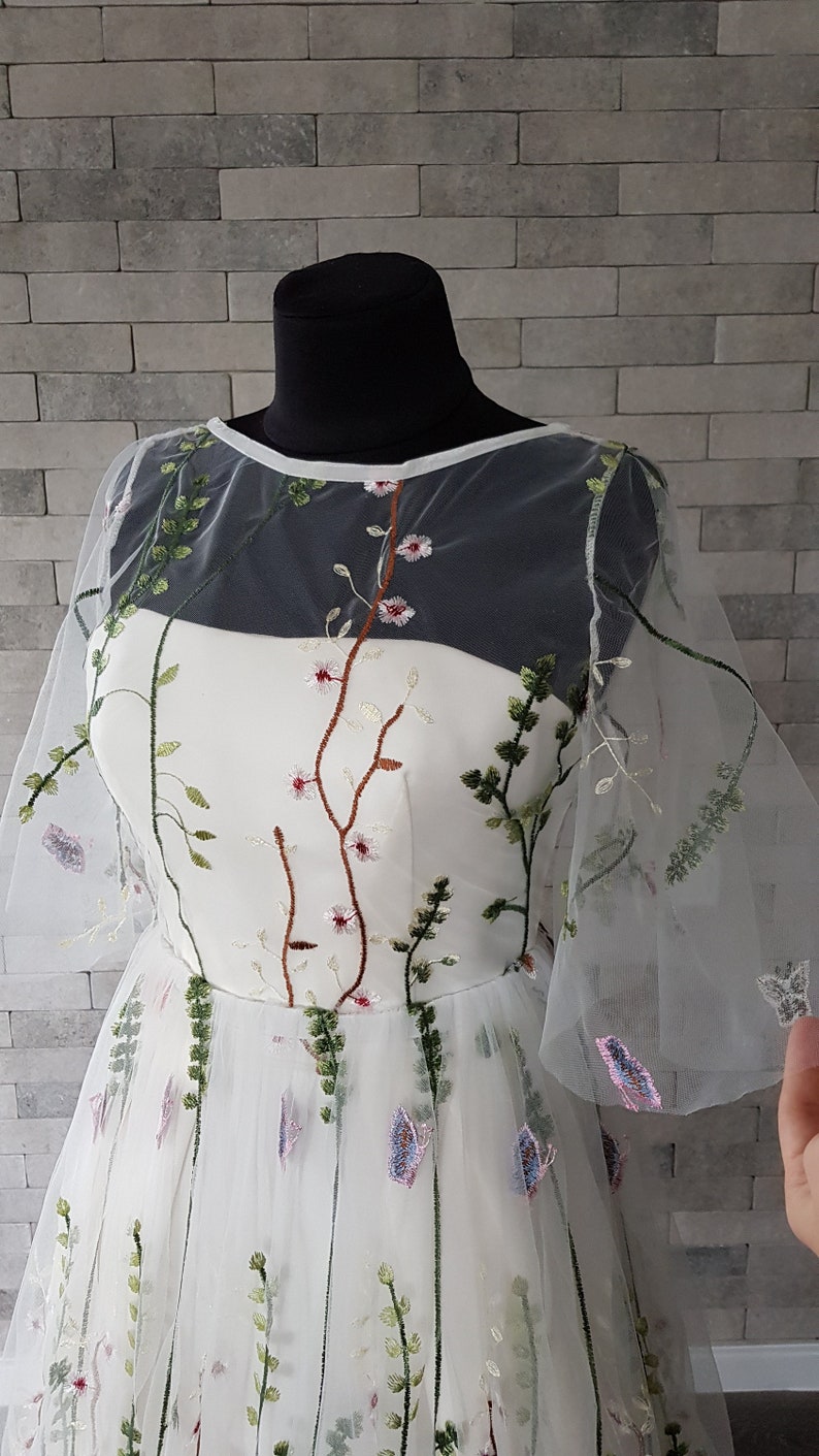 May include: A white tulle dress with floral embroidery. The dress has a sheer overlay with a round neckline and short sleeves. The embroidery features green leaves, pink flowers, and brown stems.