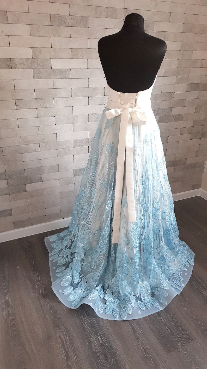May include: A light blue floor length gown with embroidered floral details. The dress features a sweetheart neckline and a white satin sash. The back of the dress is shown, revealing a corset-style back closure. The dress is displayed on a mannequin against a textured wall.