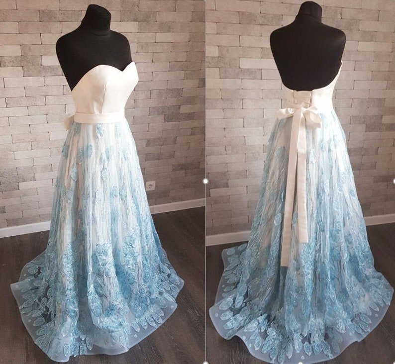 May include: Two light blue lace gowns displayed on mannequins. One gown is strapless with a sweetheart neckline and a white bodice. The other gown is backless with a white ribbon sash. Both gowns have a light blue lace overlay and a white underlay. The gowns are shown against a textured white brick wall.