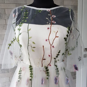 May include: A white tulle dress with floral embroidery. The dress has a sheer overlay with a round neckline and short sleeves. The embroidery features green leaves, pink flowers, and brown stems.