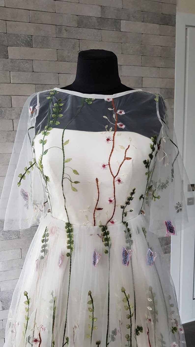 May include: A white tulle dress with floral embroidery. The dress has a sheer overlay with a round neckline and short sleeves. The embroidery features green leaves, pink flowers, and brown stems.