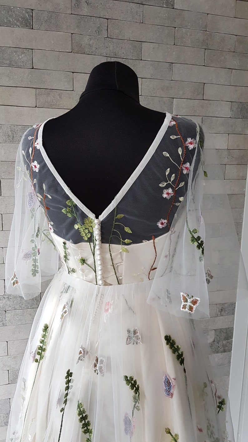 May include: White tulle dress with floral embroidery and a V-neckline. The dress has a fitted bodice and a full skirt. The embroidery features butterflies, flowers, and leaves in shades of green, pink, and white.