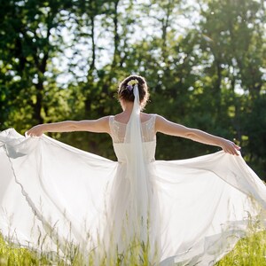 Op de afbeelding: Een bruid in een witte trouwjurk met een sluier en bloemen in haar haar staat in een veld met hoog gras en een bos op de achtergrond.