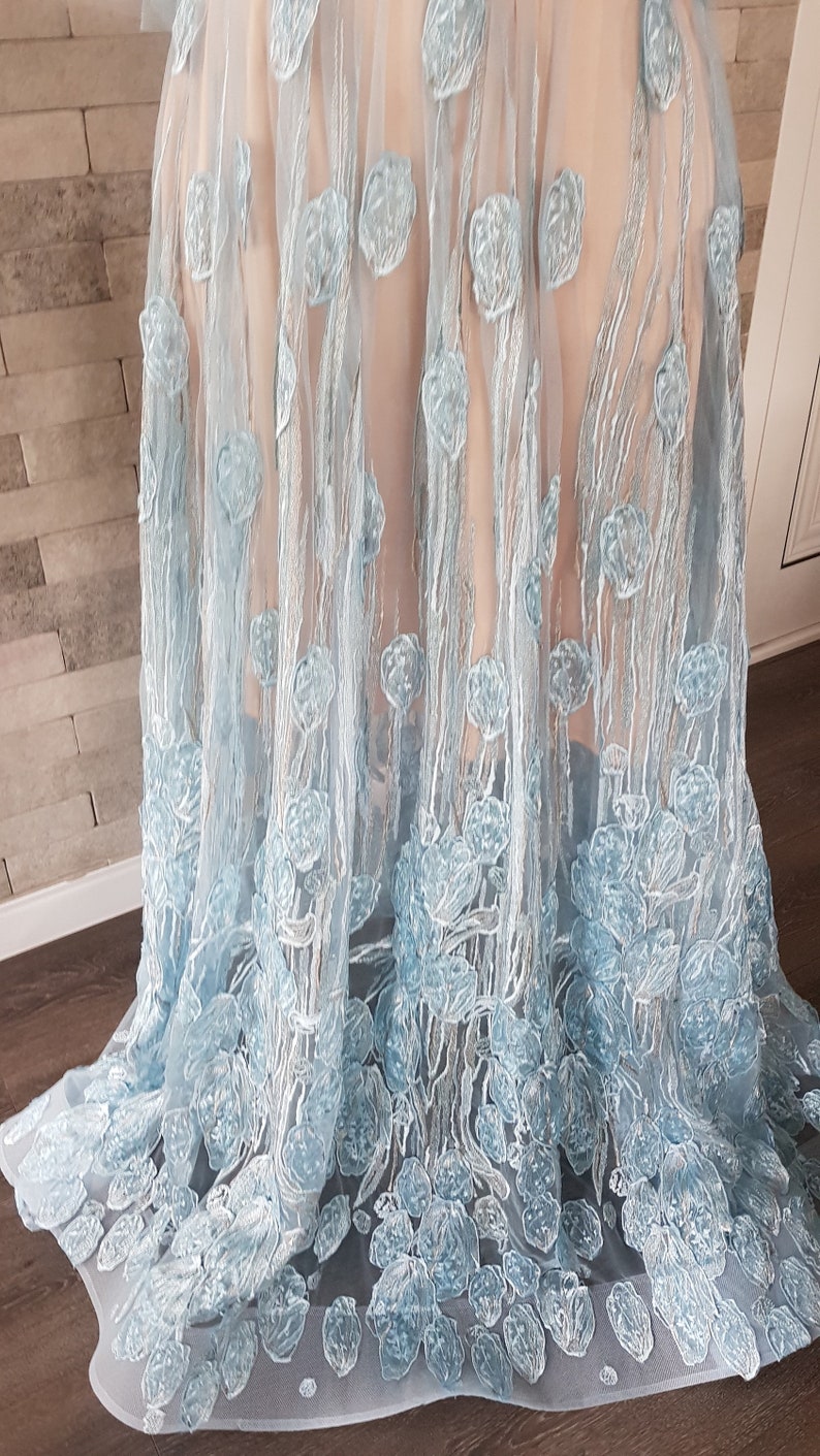 May include: A light blue tulle maxi dress features delicate floral embroidery. The embroidery is in a lighter shade of blue, creating a subtle contrast against the dress's base color. The dress is made of a sheer fabric, allowing for a glimpse of the underlayer. The skirt is full and flows gracefully, showcasing the intricate detailing of the embroidery. The dress is perfect for special occasions, offering a touch of elegance and sophistication.