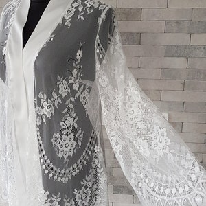 May include: White lace bridal robe with long sleeves and a satin collar. The robe is sheer with floral lace detailing throughout.  The lace has a scalloped edge. The robe is shown on a mannequin against a gray brick background. This elegant, long-sleeved lace kimono robe is perfect for bridal preparations or a luxurious addition to any wardrobe.