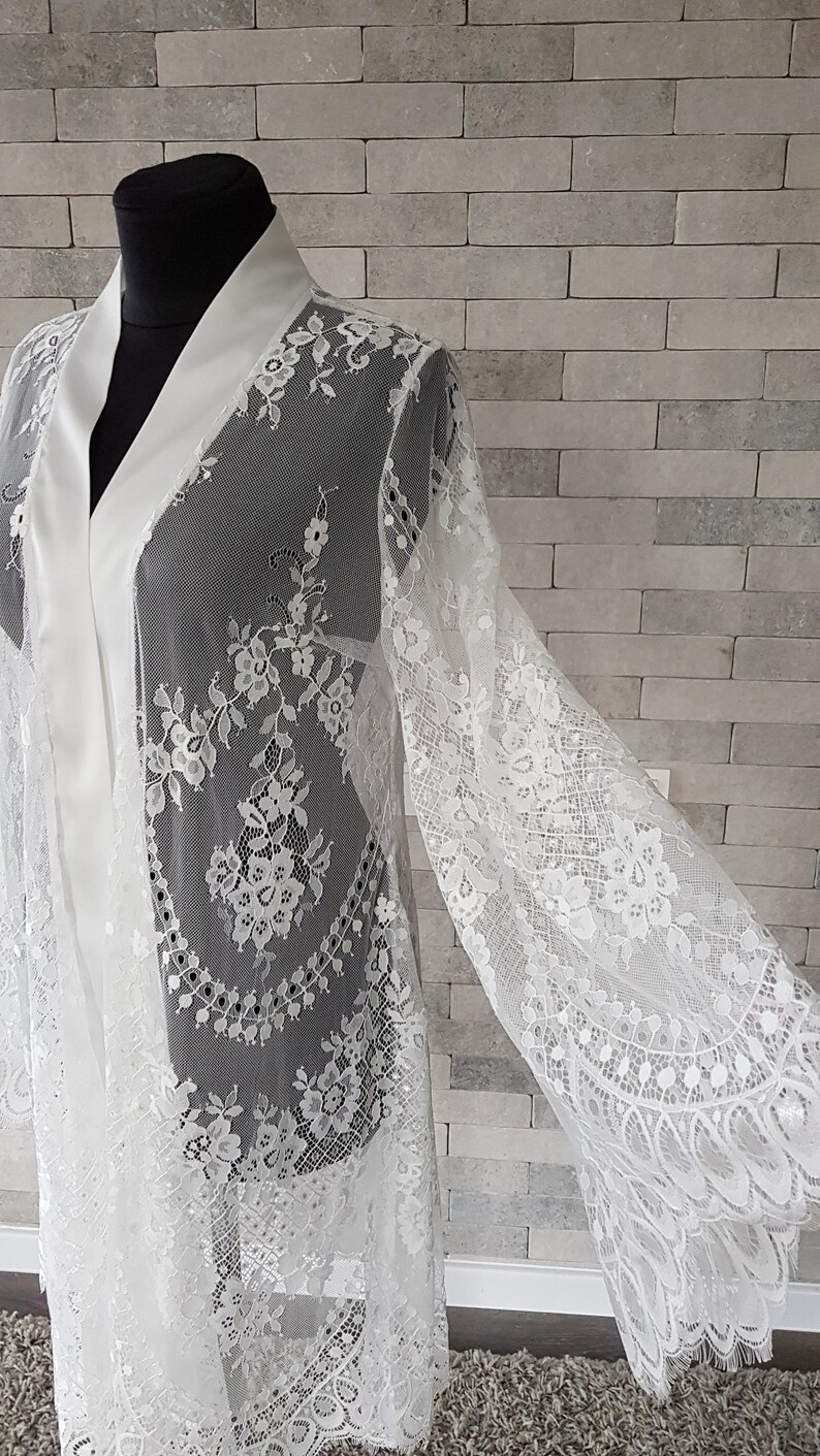 May include: White lace bridal robe with long sleeves and a satin collar. The robe is sheer with floral lace detailing throughout.  The lace has a scalloped edge. The robe is shown on a mannequin against a gray brick background. This elegant, long-sleeved lace kimono robe is perfect for bridal preparations or a luxurious addition to any wardrobe.
