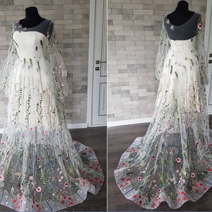 May include: A white tulle gown with a floral embroidery design. The gown has long sleeves and a full skirt. The embroidery features green leaves, pink and white flowers, and red accents.