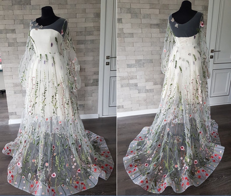 May include: A white tulle gown with a floral embroidery design. The gown has long sleeves and a full skirt. The embroidery features green leaves, pink and white flowers, and red accents.
