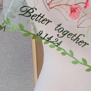 May include: White tulle fabric with green and red embroidered floral designs. The fabric has the text "Better together 9.14.24" embroidered on it.