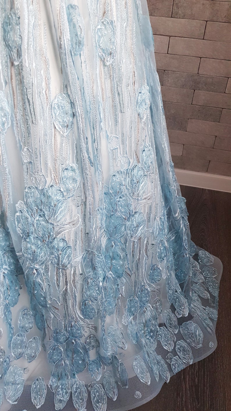 May include: Close-up of a light blue tulle gown with embroidered floral details. The fabric is sheer, and the embroidery is textured and dimensional, creating a three-dimensional effect. The light blue color is soft and delicate, and the overall design is elegant and sophisticated. The skirt of the gown is full and flowing, and the embroidery is concentrated along the bottom hemline.