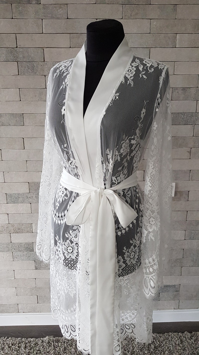 May include: White lace robe with satin trim and tie. The robe is long-sleeved and features a delicate floral lace pattern.  The satin fabric is off-white and creates a luxurious contrast to the sheer lace.  This bridal robe is perfect for getting ready on your wedding day.