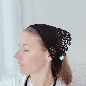 Pode incluir: Um gorro de crochê preto com um design semelhante a uma rede e lantejoulas decorativas. O gorro é usado na cabeça. A pessoa está vestindo uma camisa branca.
