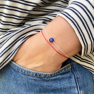 Puede incluir: Un brazalete de cuerda roja con un dije azul de ojo turco. El brazalete está en la muñeca de una persona.