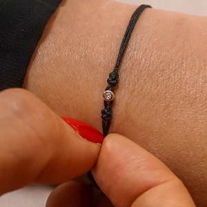 May include: A simple black string bracelet with a small, clear gemstone accent.