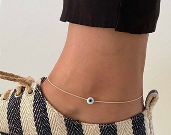 May include: A silver anklet with a blue and white evil eye charm. The anklet is on a person's ankle, and the person is wearing a striped shoe.