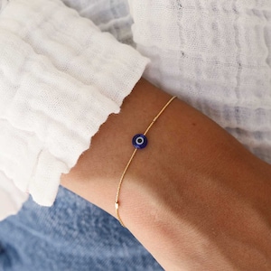 May include: A gold chain bracelet with a blue evil eye charm. The bracelet is on a person's wrist.