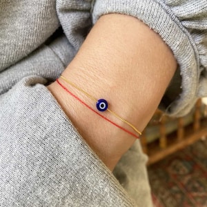 Puede incluir: Un brazalete de cadena dorada con un dije de ojo azul malvado y un brazalete de cuerda roja superpuesto.