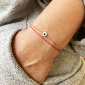 Puede incluir: Un brazalete de cadena dorada con un dije de ojo turco azul y blanco. El brazalete está superpuesto a un brazalete de cuerda roja.