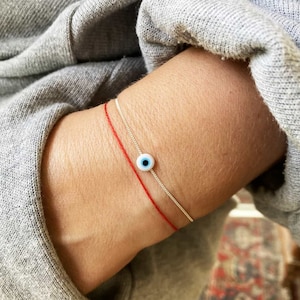 Puede incluir: Un brazalete de cadena de plata con un dije de ojo azul malvado y un brazalete de cuerda roja en la muñeca de una persona.