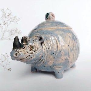 Puede incluir: Un azucarero de cerámica con forma de rinoceronte con tapa. El azucarero es de color azul claro con motas blancas y marrones. El rinoceronte tiene un cuerno negro y una cara detallada.