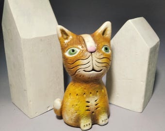 May include: A ceramic orange cat figurine with green eyes and a pink nose, featuring textured fur details and a white stripe on its forehead. The cat is positioned between two plain, white, house-shaped ceramic structures.