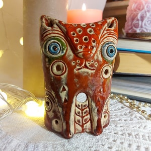 Puede incluir: Una figurita de búho de cerámica pintada a mano con un diseño caprichoso. El búho tiene ojos grandes y azules con detalles verdes, un cuerpo marrón con detalles blancos y naranjas, y un pequeño agujero en el centro de su pecho.