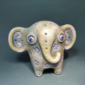 Könnte beinhalten: Eine skurrile Elefantenfigur aus Keramik in Beige-, Grün- und Blautönen. Der Elefant hat große, blaue Augen und ist mit kleinen, blauen Blumenmustern verziert. Die Ohren haben ein Wirbeldesign. Ein einzigartiger Dekorationsartikel.