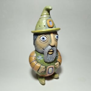 May include: A whimsical ceramic figurine of a bearded man wearing a green hat and vest. The figure has blue eyes, a gray beard, and is painted in shades of green, orange, and tan. The hat has a yellow brim and a blue and orange accent.