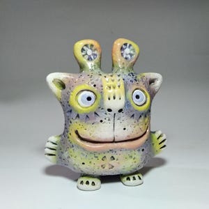 Puede incluir: Figurita de criatura de cerámica caprichosa con una cara sonriente, ojos azules grandes y orejas pequeñas. La figurita tiene una superficie texturizada con una paleta de colores amarillo, lavanda y melocotón. La criatura tiene pies y manos pequeños.